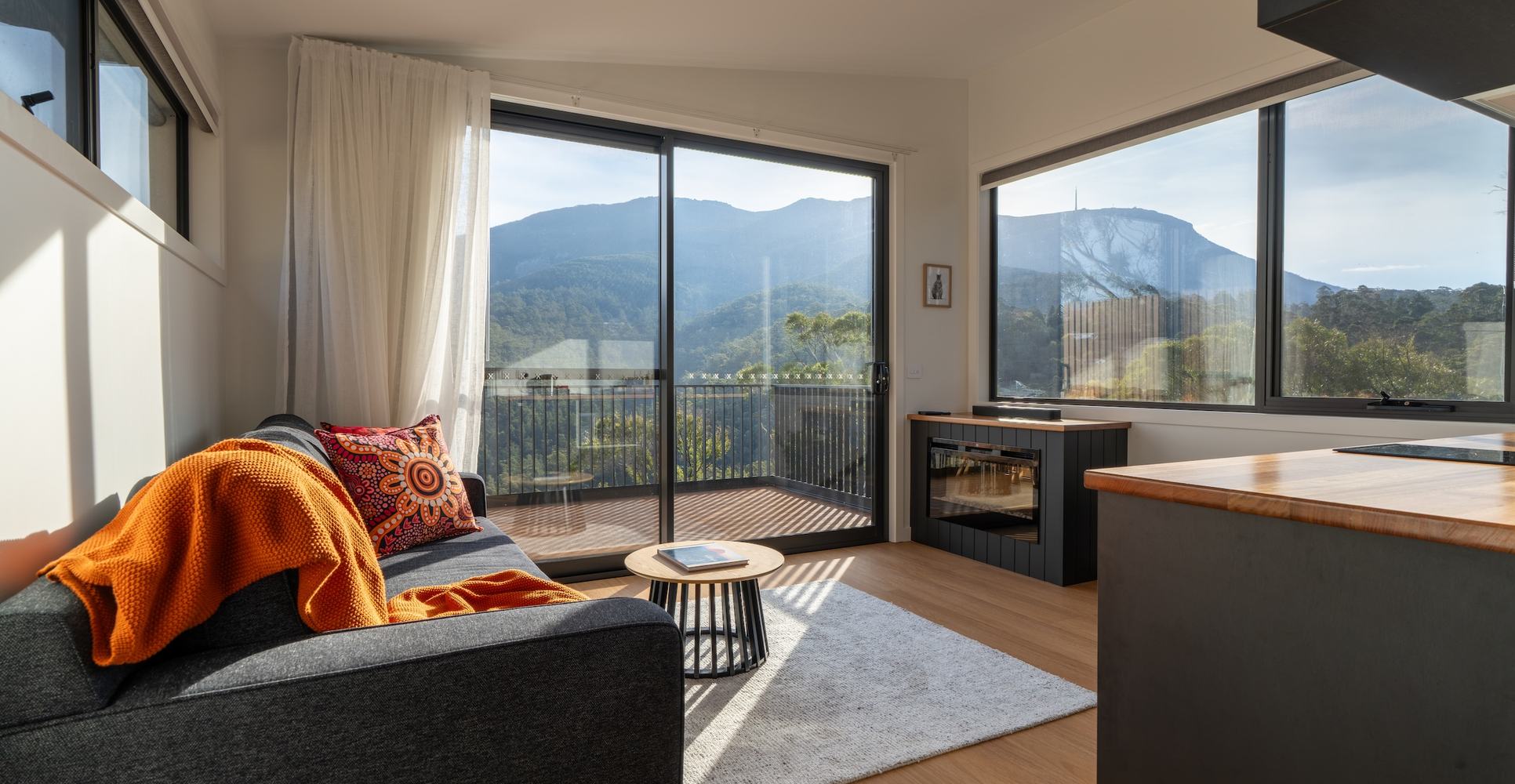 Living room of ironbark cabin with lounge, fireplace, kitchen looking out to kunanyi view