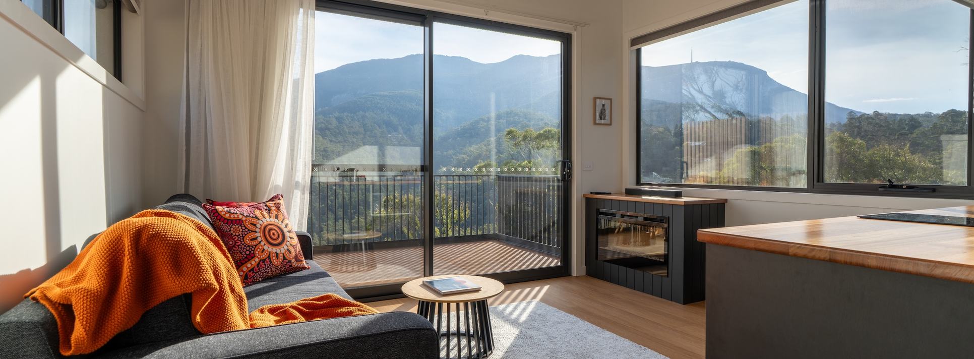 Living room of ironbark cabin with lounge, fireplace, kitchen looking out to kunanyi view